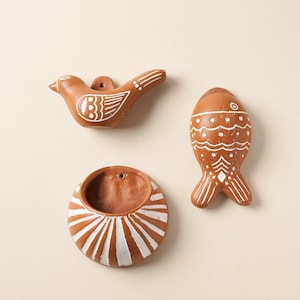 Puede incluir: Tres objetos de pared de terracota con detalles pintados en blanco. Un objeto es un pájaro, otro es un pez y el tercero es un cuenco circular con la parte posterior plana y un pequeño orificio para colgar. Acabado mate de color marrón rojizo.