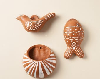 May include: Three terracotta wall-mounted objects featuring white painted details. One object is a bird, one is a fish, and one is a circular bowl with a flat back and a small hole for hanging. Each piece has a smooth, matte, reddish-brown finish.