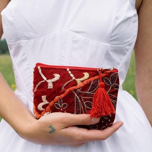 May include: A rectangular red zippered pouch featuring intricate gold embroidery, silver sequins, and a decorative orange braided trim. A matching red fabric tassel is attached to the zipper pull, adding texture to the detailed surface design.