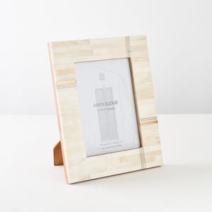 May include: A rectangular picture frame with a cream-colored tiled inlay and thin metallic accents. The frame holds a display card with the text MATR BOOMIE 5" X 7" FRAME, Designed in Austin, TX, Handcrafted in India. It features a wooden stand.
