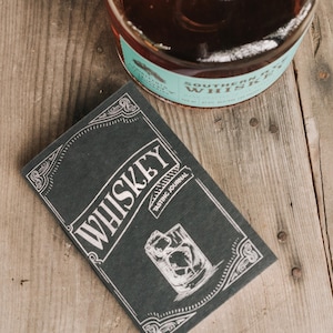 Könnte beinhalten: Ein rechteckiges, schwarzes Whiskey-Verkostungsjournal mit weißen Linienzeichnungen, dem Text WHISKEY TASTING JOURNAL und einer Abbildung eines Glases mit Eiswürfeln, platziert neben einer Glasflasche mit bernsteinfarbener Flüssigkeit und einem türkisfarbenen Etikett.