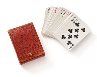 Könnte beinhalten: Ein braunes Lederetui für Spielkarten mit geprägtem Blumenmuster und einem kleinen Metallknopfverschluss, platziert neben einem aufgefächerten Stapel Standard-Spielkarten mit schwarzen Kreuz-Symbolen und der Zahl Acht auf der obersten Karte.