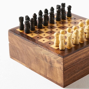 May include: A wooden chess set with a dark brown, hinged box. The chessboard is inlaid with light and dark squares. Black and white chess pieces are arranged on the board, ready for play. The set is compact and appears to be portable.