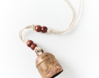 May include: A rustic, weathered brass bell with a conical shape, suspended from a light-colored, braided cord. The cord is adorned with four small, dark brown wooden beads, arranged in two pairs, providing a contrast to the metallic texture.