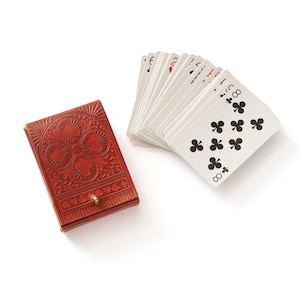 Può includere: Un astuccio per carte da gioco in pelle rossa con un motivo floreale in rilievo e una chiusura color oro, accanto a un mazzo di carte a ventaglio. Le carte mostrano il numero otto e il seme di fiori.