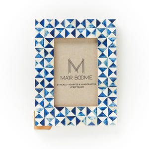 May include: A rectangular picture frame featuring a geometric pattern of alternating blue and white triangles. The center insert displays the text M MAIR BOOMIE ETHICALLY SOURCED HANDCRAFTED 4"X6" FRAME. A small brown tab is visible at the bottom left corner.