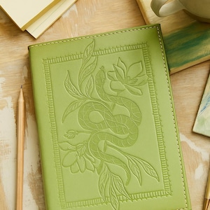 May include: A light green notebook with a rectangular embossed border and a central design featuring a snake coiled around a flower with leaves. The cover has visible stitching along the edges and a smooth, matte finish on the front surface.