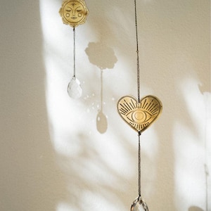 May include: A hanging mobile featuring two gold-toned metal charms suspended by thin chains. One charm is a circular sun face, and the other is a heart shape with an eye design. Each charm is attached to a clear, faceted crystal drop at the bottom.