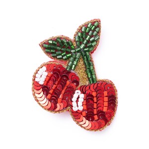 May include: A cherry-shaped brooch featuring two round fruits covered in red sequins with white bead highlights. The stems and leaves are crafted from green glass bugle beads, and the entire piece is outlined with gold metallic thread.