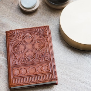 Könnte beinhalten: Ein rechteckiges Notizbuch aus braunem Leder mit einer geprägten Verzierung auf dem Vorderdeckel. Der Deckel zeigt ein zentrales blumenartiges Muster aus vier kreisförmigen Motiven mit strahlenförmigen Linien über einer Reihe von Mondphasen.