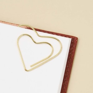 May include: A gold-toned metal paperclip shaped like a heart, attached to the top corner of a stack of white paper pages. The paperclip is constructed from a single continuous wire bent into a heart outline, providing a functional and decorative clip.