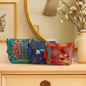May include: Three rectangular zippered pouches in teal, navy blue, and red. Each pouch features intricate embroidered floral and paisley patterns in contrasting colors, a matching fabric tassel attached to the zipper pull, and a structured shape.