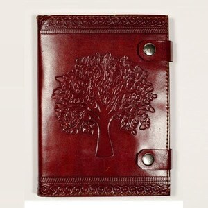 Könnte beinhalten: Ein rechteckiges Notizbuch aus dunkelbraunem Leder mit einem geprägten Baummotiv auf dem Vorderdeckel. Das Notizbuch wird durch zwei horizontale Lederriemen verschlossen, die jeweils mit einem silberfarbenen Metalldruckknopf am rechten Rand befestigt sind.