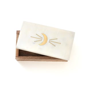 Puede incluir: Una caja rectangular blanca con un diseño de media luna dorada y rayos de sol en la tapa. La caja está abierta, revelando un interior de madera. La tapa está ligeramente entreabierta.