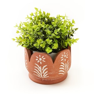 May include: A terracotta-colored planter with a scalloped top edge and white hand-painted floral designs featuring a central flower and leaf patterns. The planter holds a dense, bushy artificial plant with small, vibrant light green leaves.