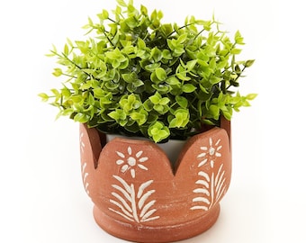 Könnte beinhalten: Ein terrakottafarbener Übertopf mit gewelltem Rand und weißen, handgemalten Blumenmustern, die eine zentrale Blüte und Blattmotive zeigen. Der Übertopf enthält eine dichte, buschige Kunstpflanze mit kleinen, leuchtend hellgrünen Blättern.