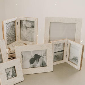 May include: A collection of rectangular and square picture frames featuring a light-colored, inlaid chevron pattern. The frames are arranged in various orientations, including hinged double-panel designs, displaying black and white photographs.