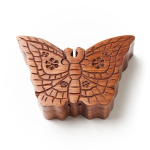 May include: A brown wooden box shaped like a butterfly with carved wing details. The surface features etched lines forming a wing pattern and small flower motifs on each side. The lid is divided into two sections that meet at the center.