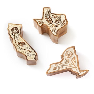 May include: Three wooden boxes shaped like the states of California, Texas, and New York. Each box features a light-colored top surface with a dark brown engraved floral design, including poppies on California, bluebonnets on Texas, and roses on New York.