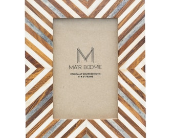 May include: A rectangular picture frame featuring a chevron pattern made of alternating strips of brown wood, white bone, and grey resin. The center insert displays the text MATR BOOMIE ETHICALLY SOURCED BONE 4" X 6" FRAME. The frame has a vertical orientation.