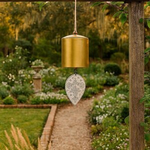 May include: A cylindrical gold-colored metal wind chime hanging from a jute rope. Attached to the bottom of the bell is a white, leaf-shaped metal wind catcher featuring a detailed black floral pattern and a symmetrical decorative design.