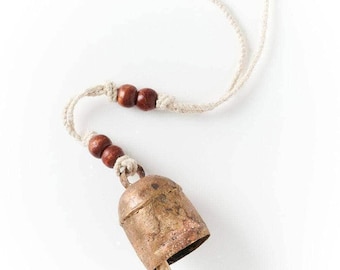 May include: A rustic, weathered copper-toned metal bell suspended from a light-colored, braided cord. The cord features four dark brown, spherical wooden beads arranged in two pairs, secured by knots, providing a decorative accent to the bell.