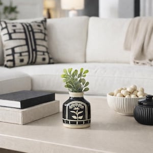 May include: A small, round ceramic vase with a black glaze and a cream-colored base. The vase features a hand-painted white floral design on the front and contains a small sprig of green artificial leaves with rounded foliage.