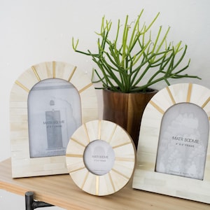 May include: Three decorative photo frames in ivory and gold tones, arranged on a wooden surface. The frames have arched and circular shapes, with a gold accent. A potted green plant sits behind the frames. The frames are labeled "MATR BODMIE 4" x 6" FRAME".