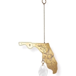 May include: A brass-colored Florida state-shaped ornament with floral and fruit engravings. A clear, teardrop-shaped crystal hangs below. The ornament is suspended by a chain and ring.