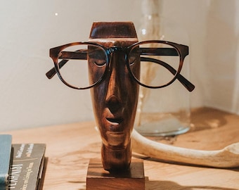 Puede incluir: Un soporte para gafas de madera de color marrón oscuro tallado con forma de rostro humano y base cuadrada. La escultura tiene un acabado liso y pulido, y sostiene un par de gafas con patrón de carey oscuro sobre el puente de la nariz.