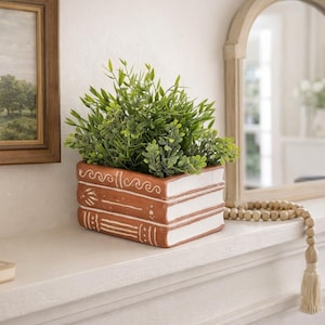 May include: A terracotta-colored ceramic planter shaped like a stack of three books with white painted details on the spines and edges. The planter holds a lush, vibrant green artificial plant with small, pointed leaves, creating a decorative display.