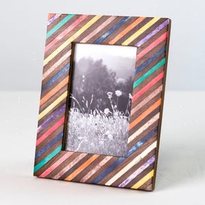 Puede incluir: Un marco de fotos rectangular con un patrón de rayas diagonales de listones de madera marrón oscuro intercalados con tiras de resina de colores vibrantes en tonos turquesa, rosa, naranja, amarillo, morado y azul, con una foto en blanco y negro.