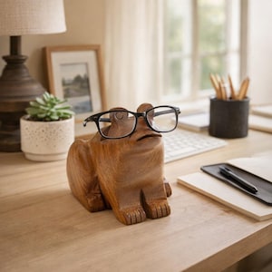 Op de afbeelding: Een houten kikker-bureaubladorganizer met een bril met zwart montuur. De kikker is bruin met uitgesneden details en de bril zit op zijn hoofd. De organizer staat op een houten bureau.
