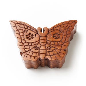 May include: A wooden butterfly-shaped box with intricate carved details. The box is made of a warm brown wood and features floral and geometric patterns. The wings are separated, suggesting a hinged design. The box is on a white background.