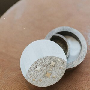 Puede incluir: Una caja redonda de madera con acabado bicolor. La tapa presenta una sección pintada de blanco y una sección de madera natural incrustada con pequeñas estrellas y puntos de latón. La base es un anillo de madera hueco a juego.