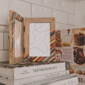 May include: A hinged double photo frame made of light brown wood, featuring diagonal stripes of orange, blue, and white resin accents on the inner edges. The frame holds two white inserts with thin, abstract, light brown line drawings.