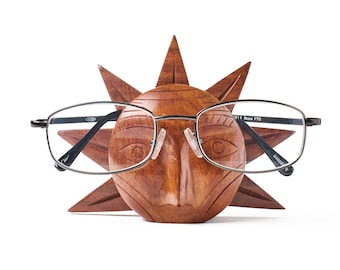 Puede incluir: Un soporte para gafas de madera tallada con forma de sol, con un rostro central y rayos triangulares. La madera marrón presenta líneas grabadas alrededor de los ojos y la nariz. Un par de gafas de metal de montura fina descansa sobre el rostro.