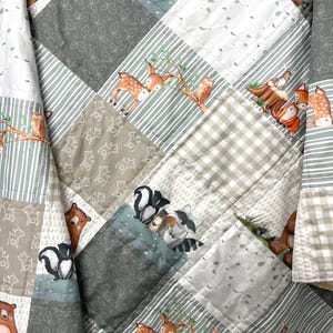 May include: A patchwork quilt featuring woodland animal illustrations. The quilt showcases squares with images of deer, bears, foxes, skunks, raccoons, and owls. The color palette includes shades of gray, white, and brown, with leaf and stripe patterns.