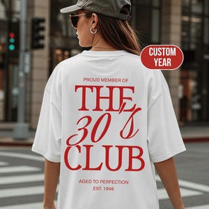 Op de afbeelding: Een wit t-shirt met rode tekst op de achterkant die luidt PROUD MEMBER OF THE 30'S CLUB, AGED TO PERFECTION, EST. 1996. Het shirt wordt gedragen met een olijfgroene baseballpet en een zonnebril en heeft een casual ontwerp met korte mouwen.