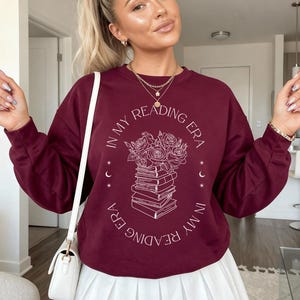Könnte beinhalten: Ein kastanienbraunes Langarm-Sweatshirt mit Rundhalsausschnitt und einer weißen Strichzeichnung eines Bücherstapels, der mit Rosen gekrönt ist. Der Text "IN MY READING ERA" ist kreisförmig um die zentrale Illustration angeordnet.