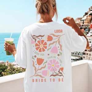 May include: White t-shirt with a floral design in orange, pink, and brown. The text on the back reads "LUISA", "I SAID YES", "FROM MS. TO MRS.", and "BRIDE TO BE". The shirt is a relaxed fit.
