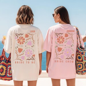 May include: Two oversized t-shirts, one natural and one light pink, with floral designs and text. The natural shirt says "Bride to Be" and "Luisa." The pink shirt says "Bride Crew" and "Johanna."