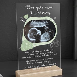 Op de afbeelding: Een helder acryl bord met een groene en witte achtergrond. Het bord heeft een zwart-witte echografie afbeelding van een baby in het midden. De tekst op het bord luidt "alles gute zum 1. vatertag", wat vertaald "alles goed voor de 1e vaderdag" betekent.