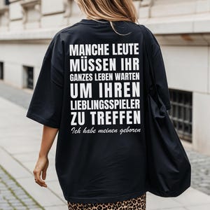 Könnte beinhalten: Ein schwarzes T-Shirt mit kurzen Ärmeln und weißem Text auf der Rückseite: MANCHE LEUTE MÜSSEN IHR GANZES LEBEN WARTEN UM IHREN LIEBLINGSSPIELER ZU TREFFEN Ich habe meinen geboren. Das T-Shirt hat eine lockere Passform und einen Rundhalsausschnitt.