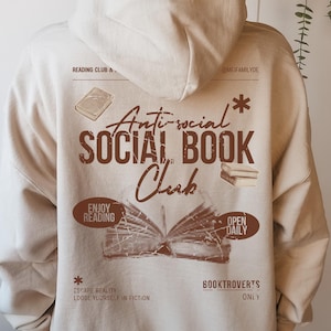 Könnte beinhalten: Beigefarbener Hoodie mit einem Aufdruck auf der Rückseite. Das Design zeigt den Text "Anti-social SOCIAL BOOK Club" in einem Vintage-Stil, Buchillustrationen und die Sätze "Enjoy Reading" und "Open Daily".