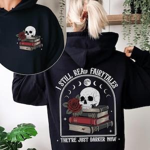 Könnte beinhalten: Schwarzer Kapuzenpullover mit Schädel- und Rosen-Design. Auf der Rückseite steht der Text "I STILL READ FAIRYTALES THEY'RE JUST DARKER NOW". Auf der Vorderseite sind ein Schädel, eine rote Rose und ein Bücherstapel abgebildet.
