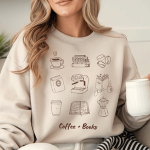Op de afbeelding: Beige sweatshirt met bruine illustraties van koffie en boeken. De tekst "Coffee + Books" is onder de tekeningen gedrukt. De persoon op de foto houdt een witte mok vast.