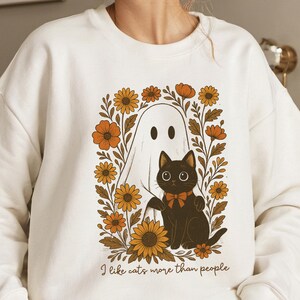 Könnte beinhalten: Weißes Sweatshirt mit einer Grafik eines Geistes und einer schwarzen Katze mit Fliege, umgeben von orangefarbenen und gelben Blumen. Der Text "I like cats more than people" ist darunter gedruckt.