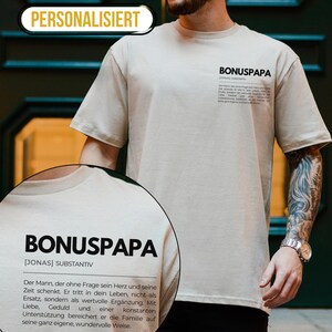 May include: A beige t-shirt with the text "BONUSPAPA [JONAS] SUBSTANTIV" and a definition of the word. The text is in black and the shirt is worn by a person with tattoos on their arm.
