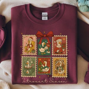 Könnte beinhalten: Burgunderfarbenes Sweatshirt mit einem skurrilen Design von Enten in festlichen Briefmarken. Die Briefmarken sind in einem Raster angeordnet und zeigen Enten in verschiedenen Urlaubsszenen, mit einer roten Schleife oben. Der Text "Introvert Season" steht unten.